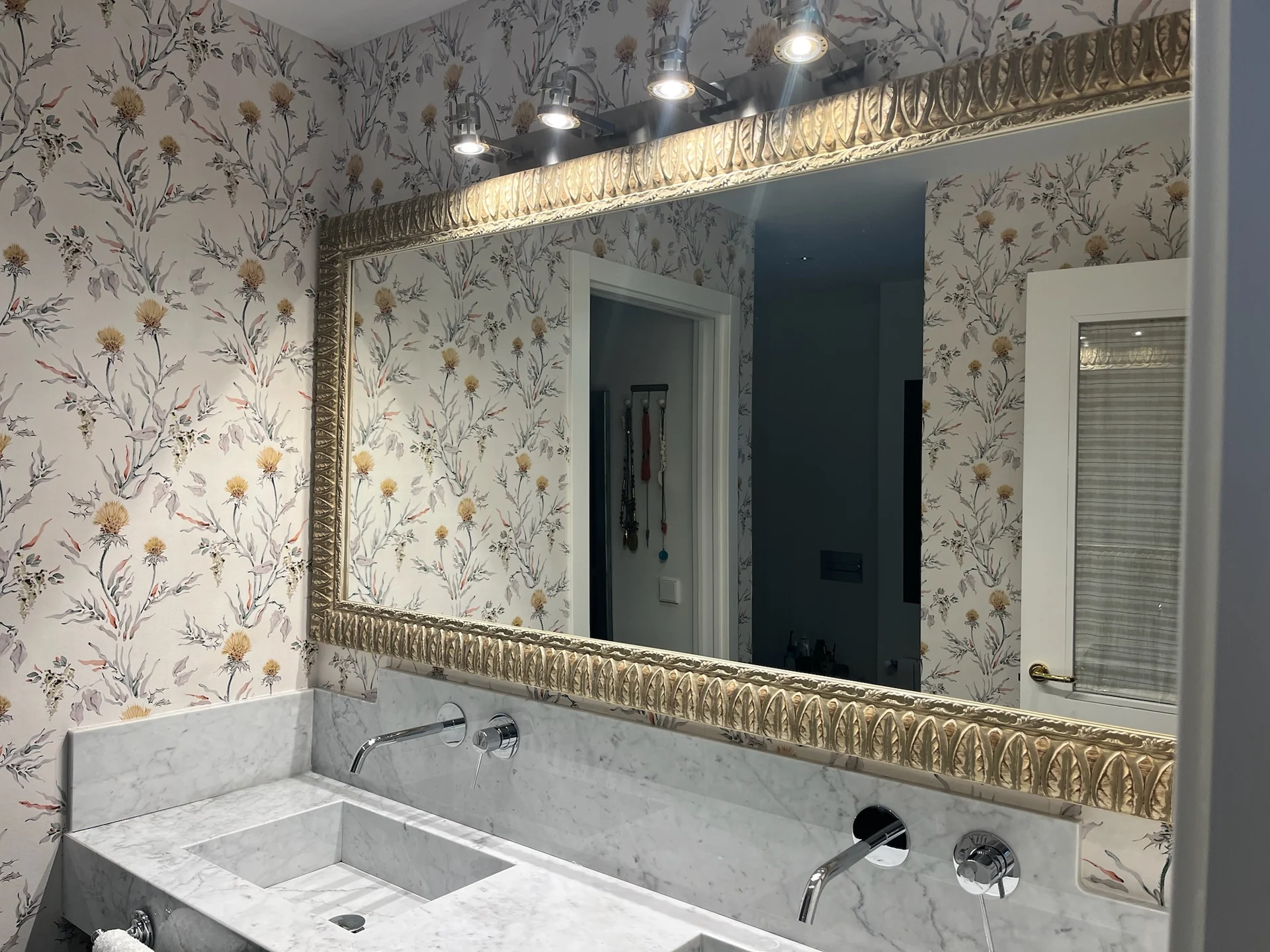 Baño con papel pintado vegetal, espejo clásico e iluminación decorativa.
